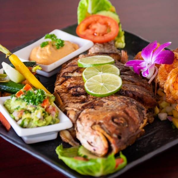 Whole fried fish with lime slices, guacamole, dipping sauce, and vegetables on a black plate.
