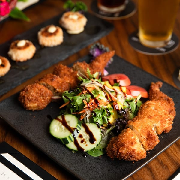Crispy fried chicken with a side salad and cucumber slices, drizzled with balsamic glaze, on a black plate.